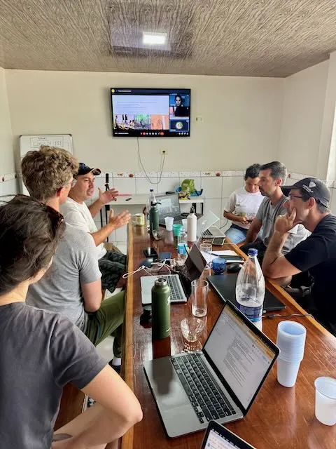 The team working around a long wooden table at the Amazon field station, laptops open, on a video call with remote colleagues
